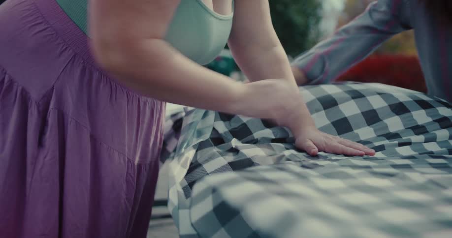 A slow-motion close-up of two women's hands smoothing a black and white checkered tablecloth onto an outdoor table