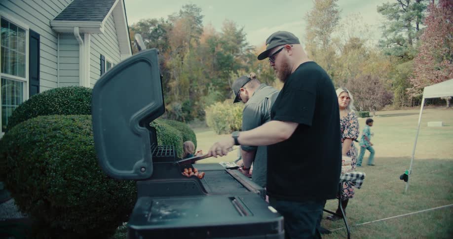 A man grills hot dogs for a backyard family cookout while children play in the background