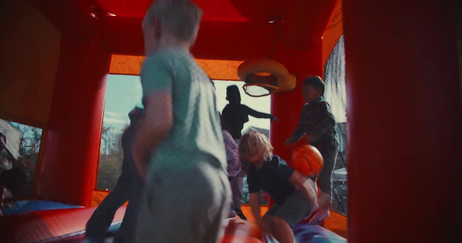 A slow-motion interior shot captures a group of diverse children laughing and joyfully jumping inside a bounce house at a sunny outdoor party