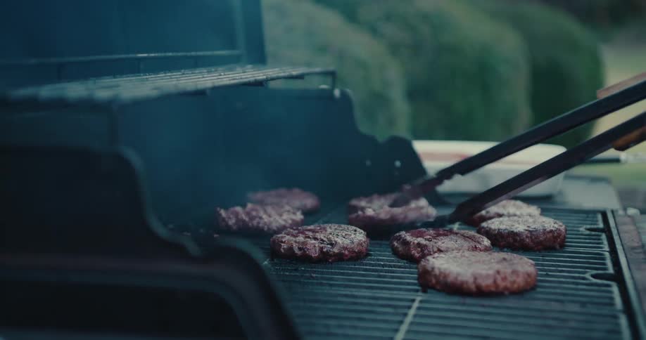 Sizzling beef patties are flipped with tongs on a smoking barbecue grill with visible flames