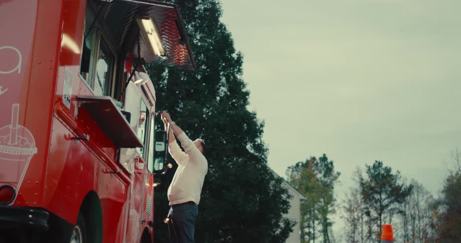 In slow motion, a man carefully hangs a string of festive lights on a red food truck