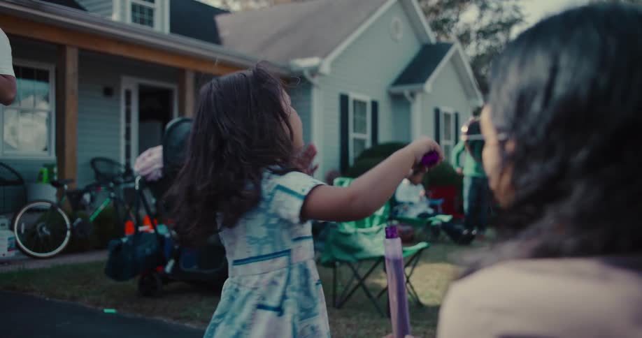 A young girl with pigtails joyfully blows bubbles in slow motion during a family gathering in the front yard