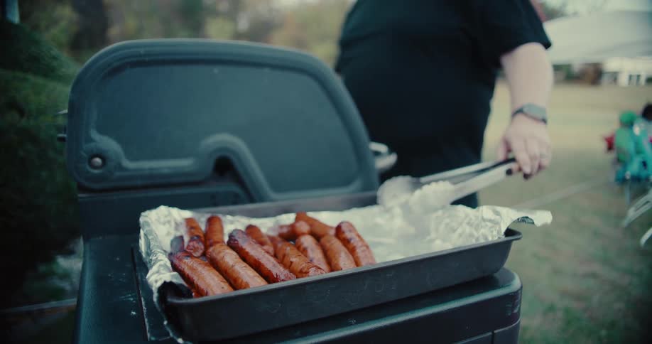 A person holds tongs while freshly grilled sausages steam in a pan next to a barbecue at an outdoor cookout