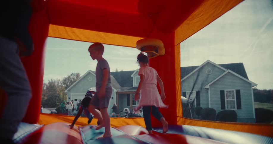 A diverse group of kids joyfully jump and play together inside a colorful bounce house during a sunny neighborhood party in slow motion