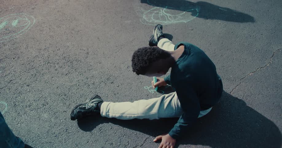 A high-angle view of a young boy sitting on the pavement drawing with sidewalk chalk as his friend watches