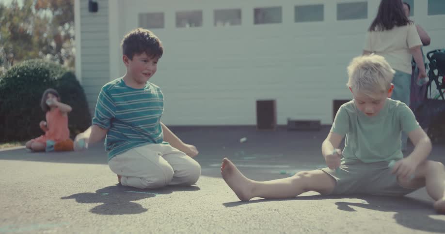 Two barefoot boys draw with sidewalk chalk on a driveway in slow motion while other kids play in the background on a sunny day