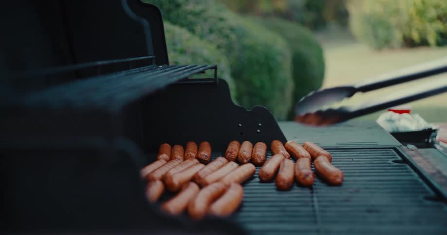 A close-up slow-motion view of a person using tongs to flip hot dogs on a backyard barbecue grill