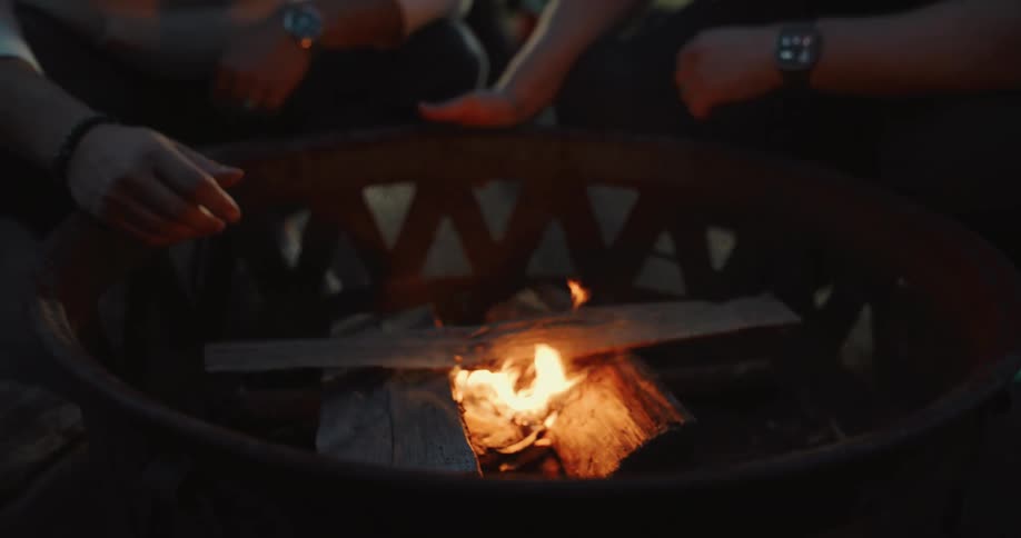 A close-up shot shows two people warming their hands over a small, crackling fire in a metal fire pit at dusk