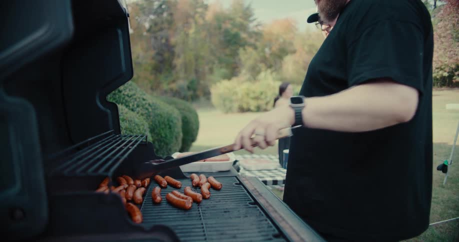A man in a black shirt and baseball cap grills small sausages on a barbecue in slow motion during a backyard gathering
