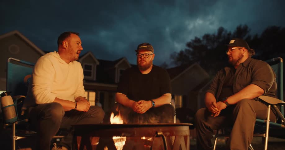Three male friends sit in camping chairs and talk around a warm fire pit at night