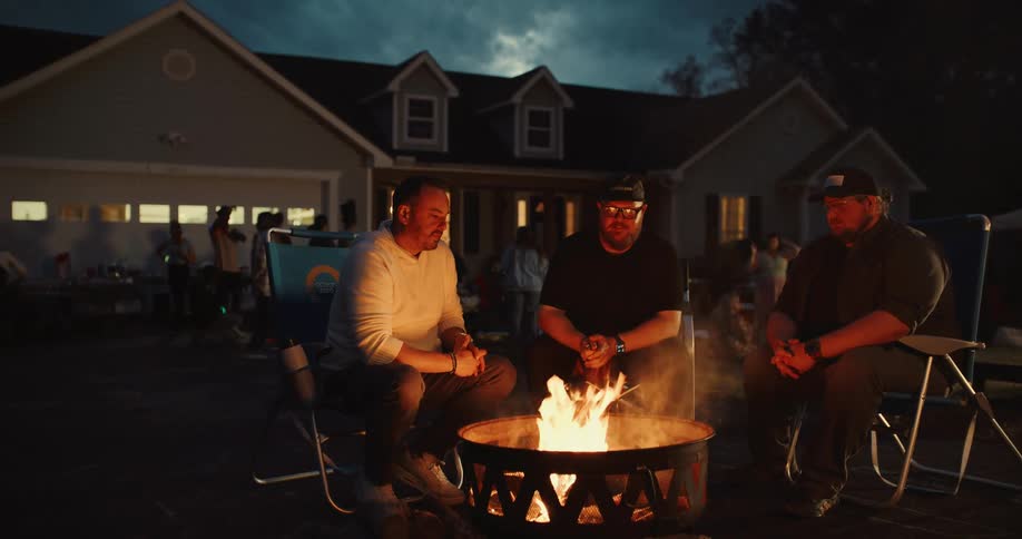Three male friends enjoy a warm conversation around a fire pit during a lively backyard gathering at dusk