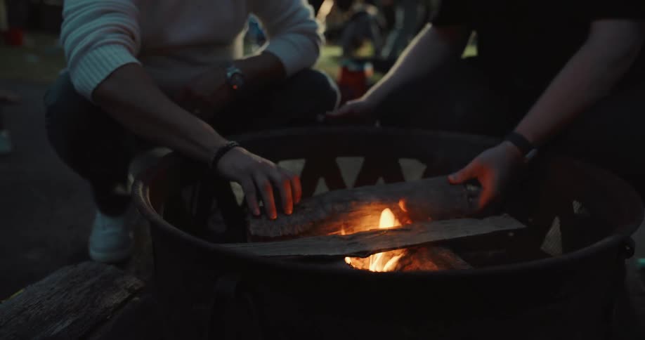 Two men carefully place a log on a budding campfire in a metal fire pit during an outdoor gathering at dusk