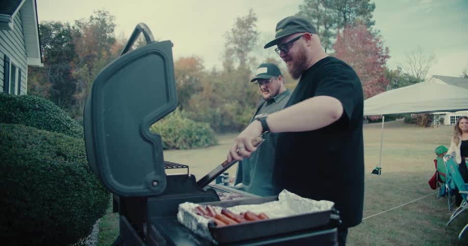 A man with a beard and glasses flips hot dogs and burgers on a grill during a casual backyard cookout with friends