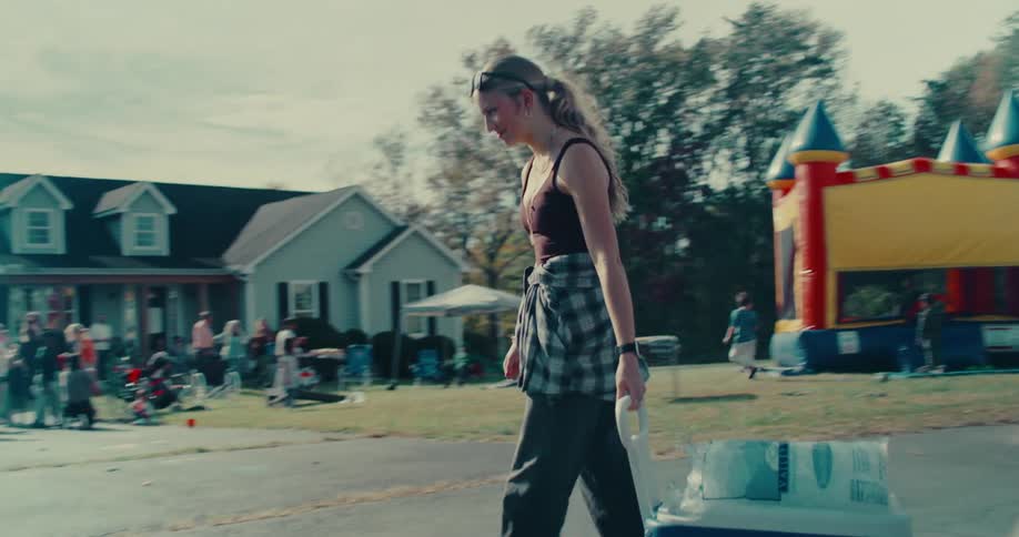 A young woman arrives at a suburban daytime party, pulling a cooler towards the crowd and bouncy house in slow motion