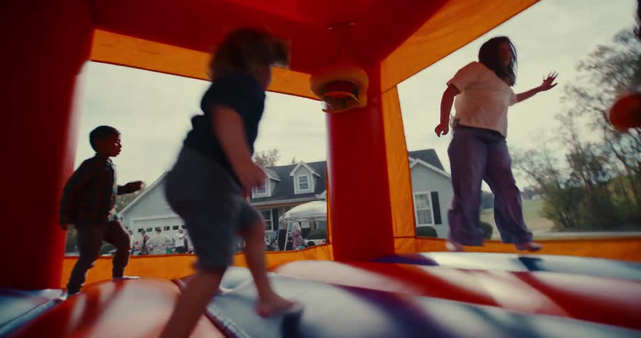 A group of happy, diverse children jump and play together in a colorful bounce house during an outdoor party, filmed in slow motion from inside