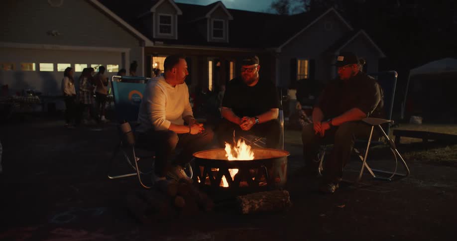 Three male friends enjoy a conversation around a warm fire pit during an evening social gathering in a driveway