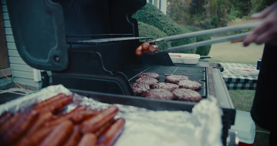Slow motion close-up of a person using tongs to grill hamburgers and hot dogs at a backyard barbecue