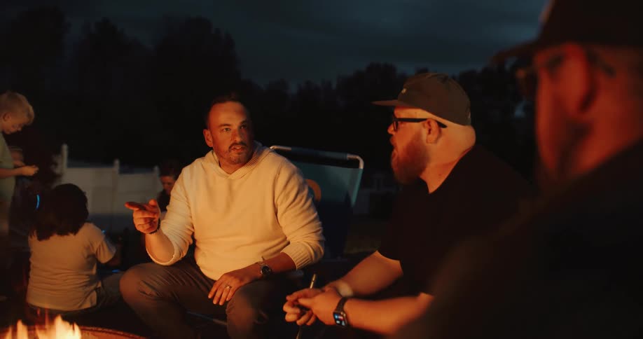 Three men have a deep conversation around a backyard bonfire at night while their children play in the background