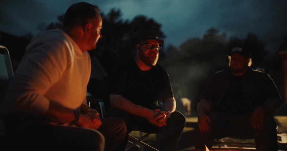 A group of male friends share stories and enjoy a conversation while sitting around a campfire at night