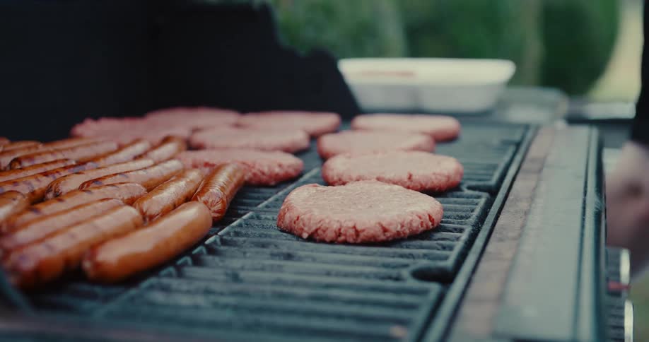 Slow motion close-up of a hand placing a raw hamburger patty on a barbecue grill alongside hot dogs