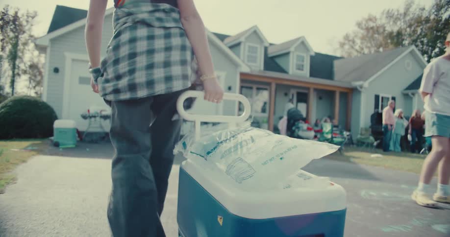 A young woman pulls a cooler with a bag of ice up a driveway to a daytime house party in slow motion