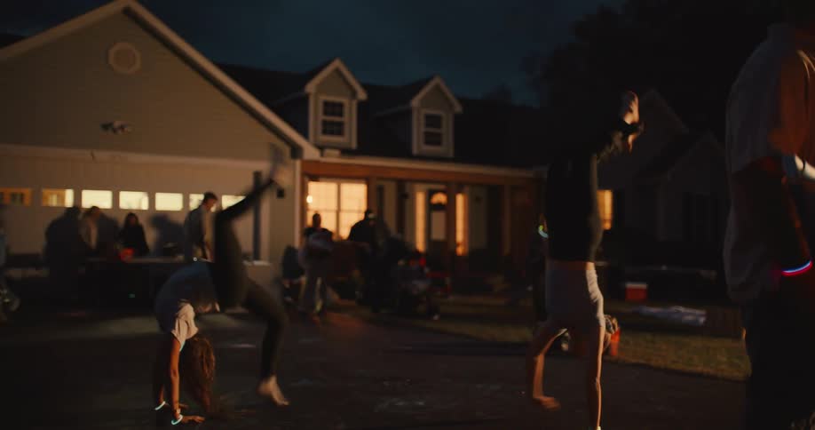 Two energetic young women do cartwheels and handstands in slow motion in their driveway during a nighttime block party