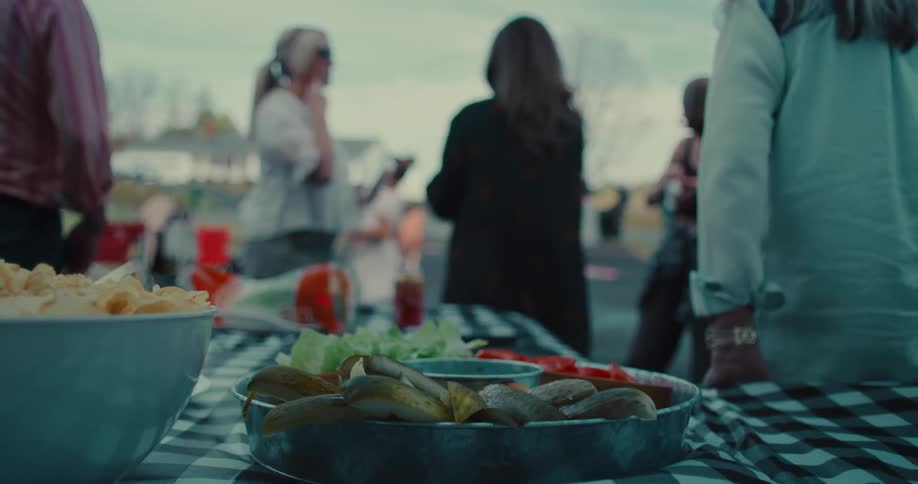 A tray of pickles, tomatoes, and lettuce sits on a checkered tablecloth with people mingling at an outdoor gathering in the background