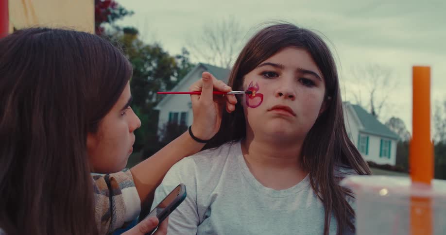 A focused teenage girl paints a colorful design on the cheek of her unimpressed friend at an outdoor event