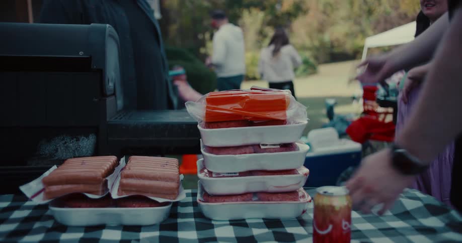 A person sets down stacks of raw hamburger patties and cheese on a checkered tablecloth at a backyard barbecue