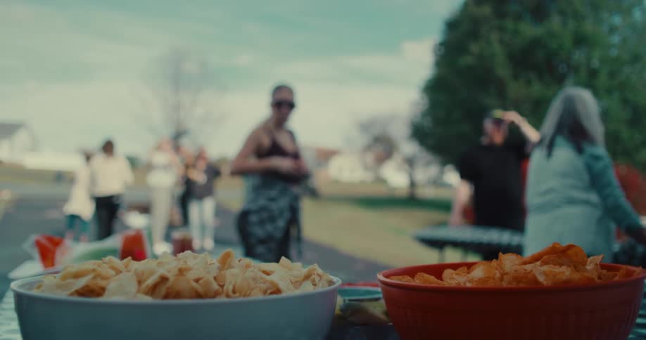 Bowls of potato chips sit on a picnic table with people mingling in the background at an outdoor party