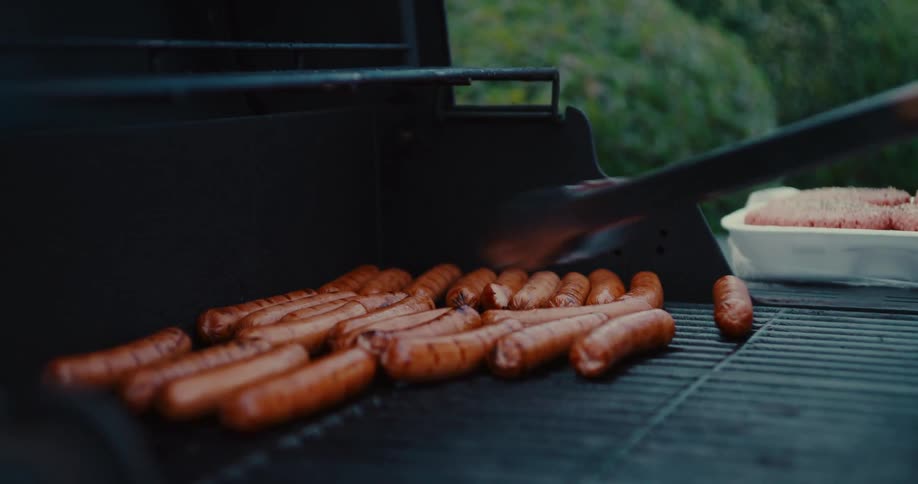 Slow motion close-up of a person turning hot dogs with tongs on a barbecue grill during a backyard cookout