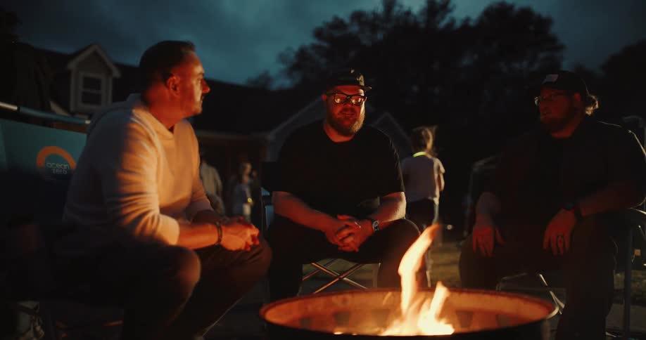Three male friends enjoy a relaxed conversation around a crackling bonfire during a neighborhood gathering at dusk