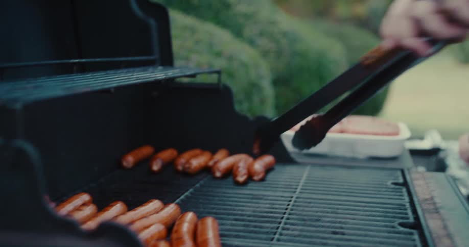 A person flips sausages with tongs while grilling them on a barbecue in slow motion