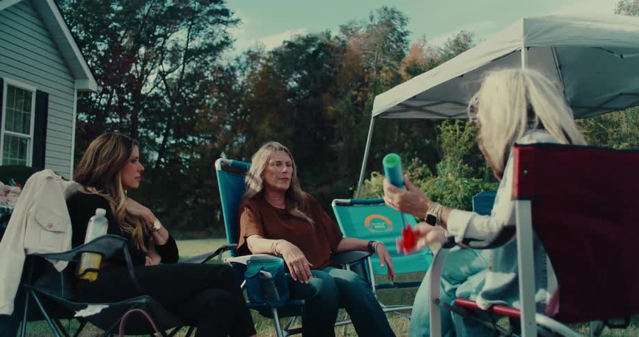 A woman with a nasal cannula has a relaxed conversation with two friends or family members while sitting in lawn chairs in a backyard