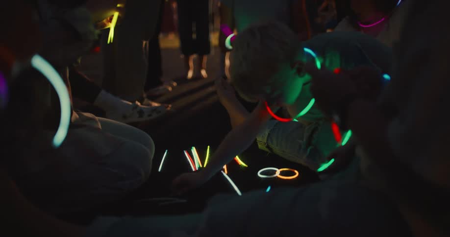 A group of happy children play with colorful glow sticks on the ground during a summer night party