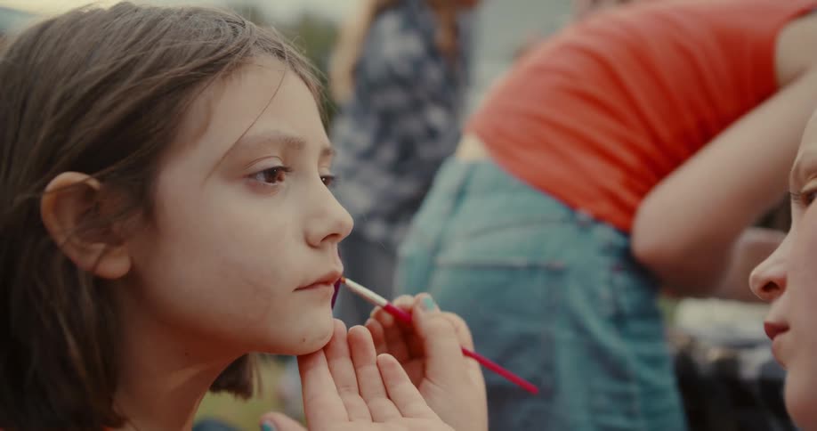 An artist carefully applies purple face paint to a young girl's lips with a small brush at an outdoor event