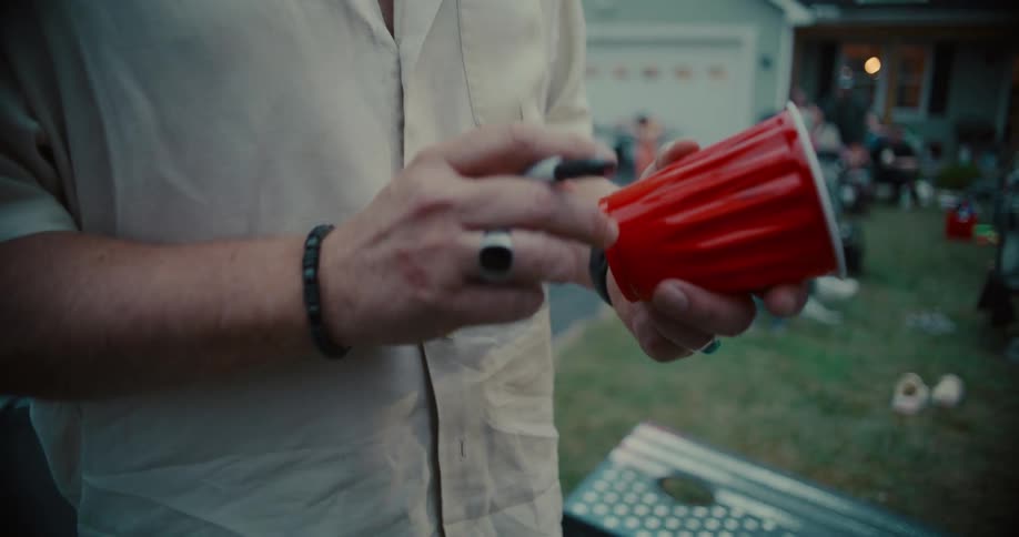 A man writes his name on a red plastic cup with a marker at an outdoor backyard party