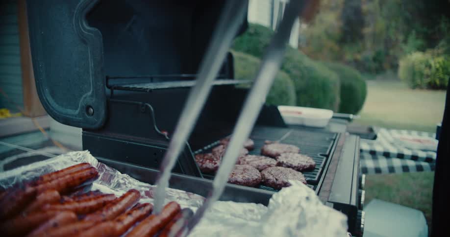 A person tends to a smoking barbecue grill, using tongs to move hot dogs alongside sizzling hamburger patties