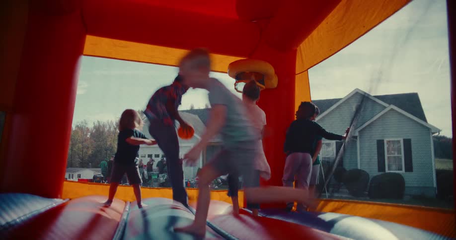 A diverse group of kids happily jump and play together inside a colorful bounce house during a sunny neighborhood party in slow motion