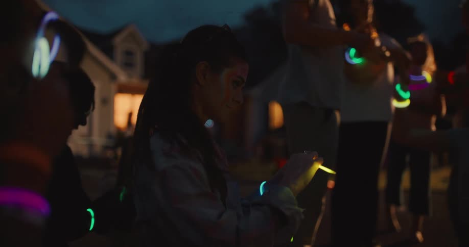 A young woman with face paint smiles as she activates a bright yellow glow stick at an outdoor night party with friends