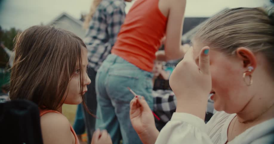 A teenage girl carefully paints a younger girl's face with a small brush at a lively outdoor gathering