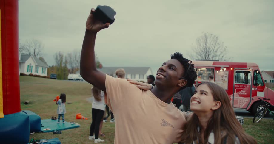 A happy young couple takes a selfie together at an outdoor community event with a food truck and other people in the background