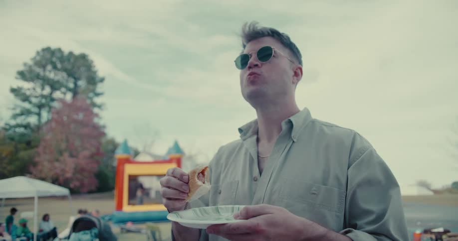 A young man in sunglasses takes a satisfying bite of a hot dog in slow motion at an outdoor party with a bouncy castle in the background