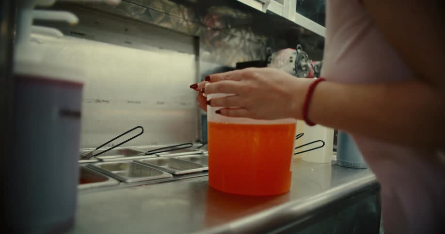 A food truck worker secures the lid on a large container of orange syrup before moving it off the stainless steel counter