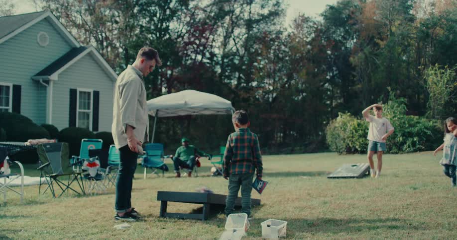 A young boy tosses a beanbag in slow motion during a game of cornhole at a backyard family gathering