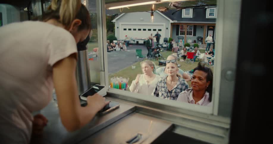 View from inside a food truck as a worker takes payment from a line of diverse customers at a lively neighborhood block party
