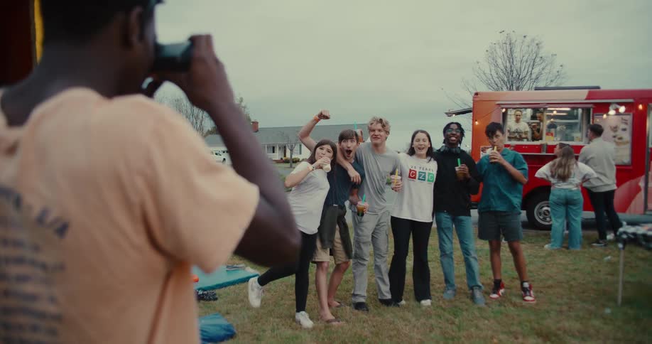 Over-the-shoulder slow-motion shot of a teenager taking a picture of his diverse group of friends posing at an outdoor party