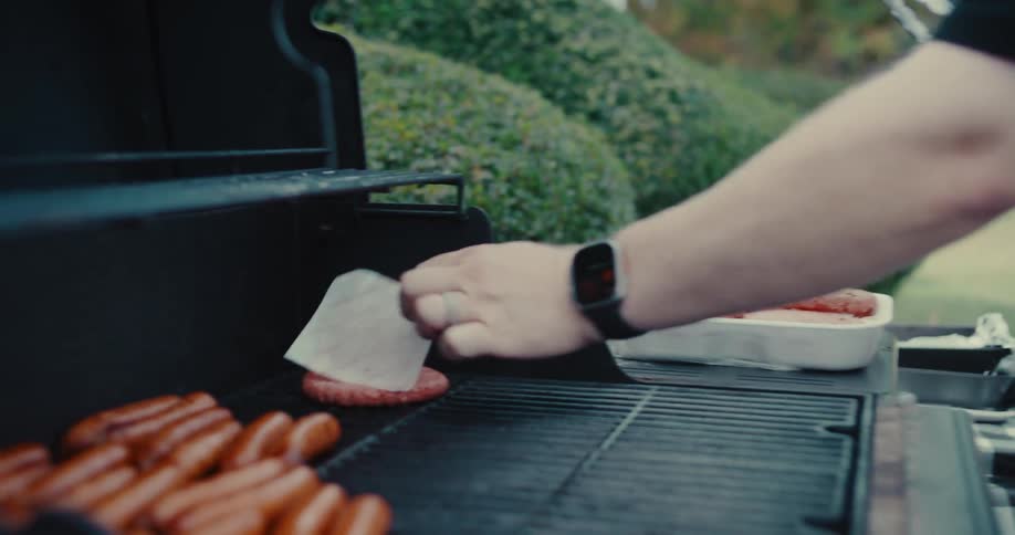 Slow motion close-up of a person placing a raw hamburger patty on a hot grill during a backyard cookout