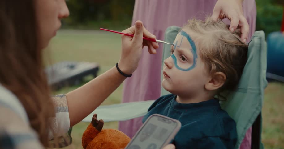 An artist carefully paints a blue superhero design on a toddler's face while sitting in a camping chair outdoors