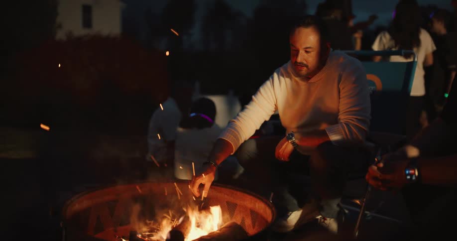 A man tends to a crackling fire pit at a backyard party at night before sitting down to talk with a friend